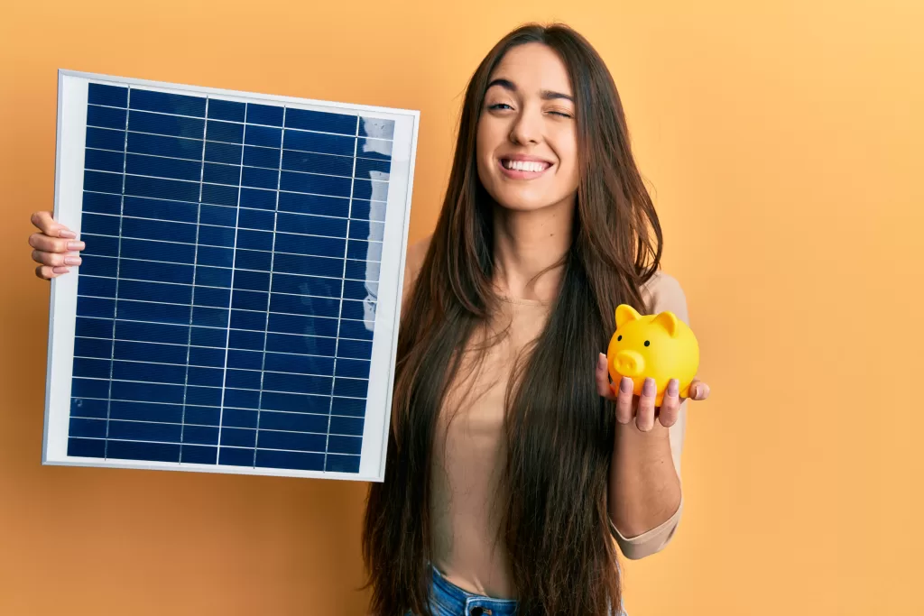 The girl is holding a solar panel in one hand and a piggy bank in the other