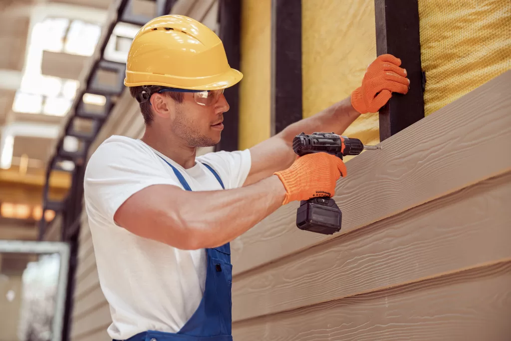 Handsome young man construction worker wearing safety helmet and work gloves while installing exterior wood siding