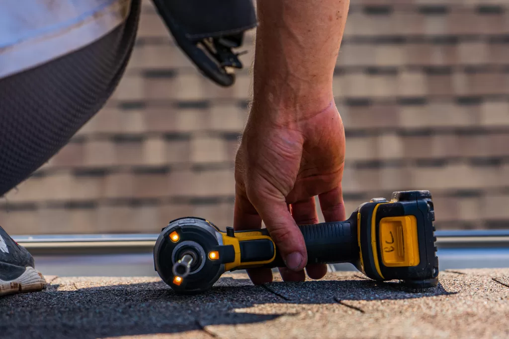 A roofer lifts a drill from the ground against a roof background
