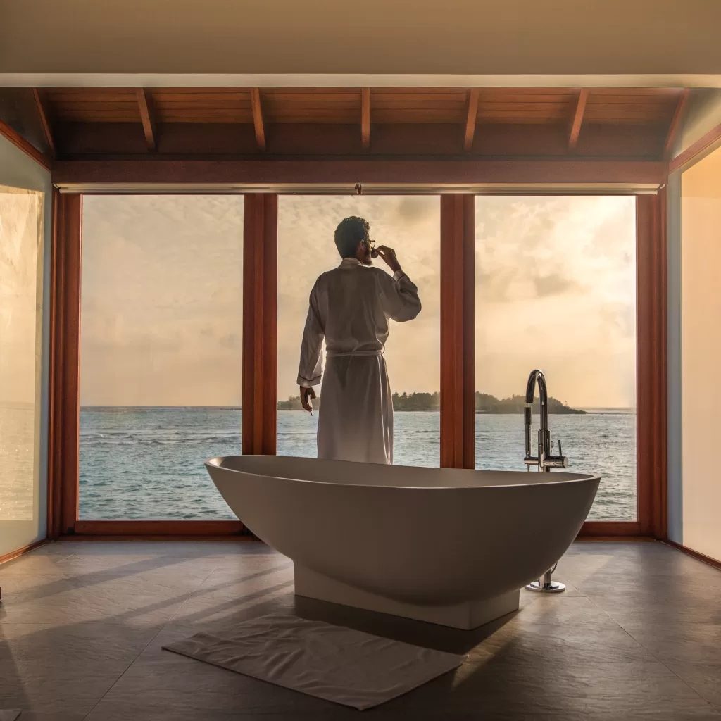 A man is relaxing in the bathroom, sipping wine with a sunset background