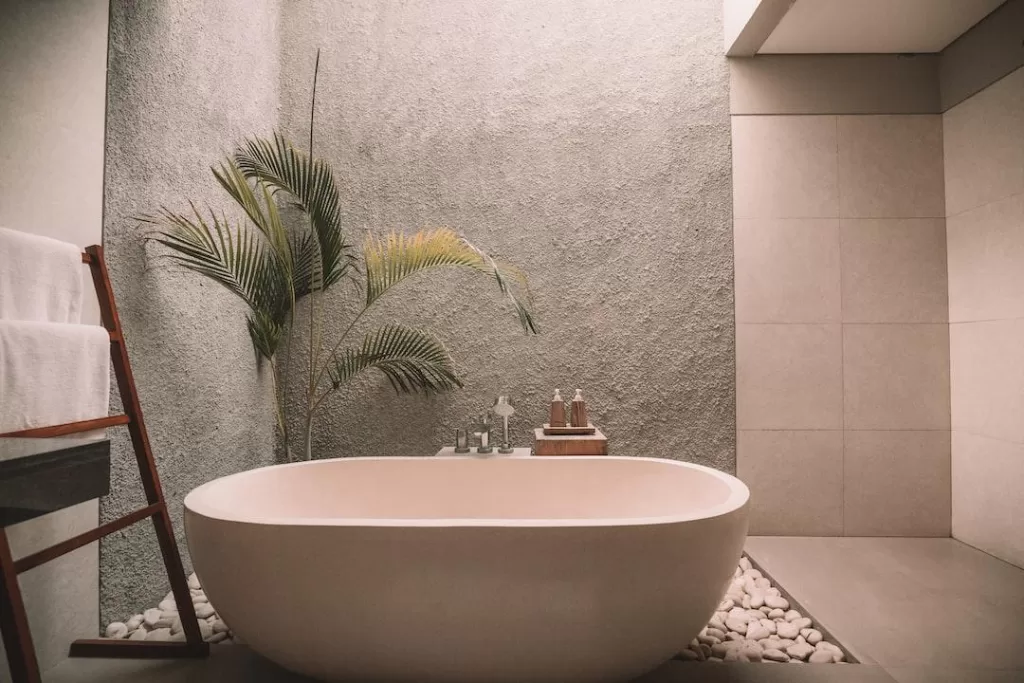 A freestanding beige tub sits on a bed of decorative rocks beside a large houseplant.