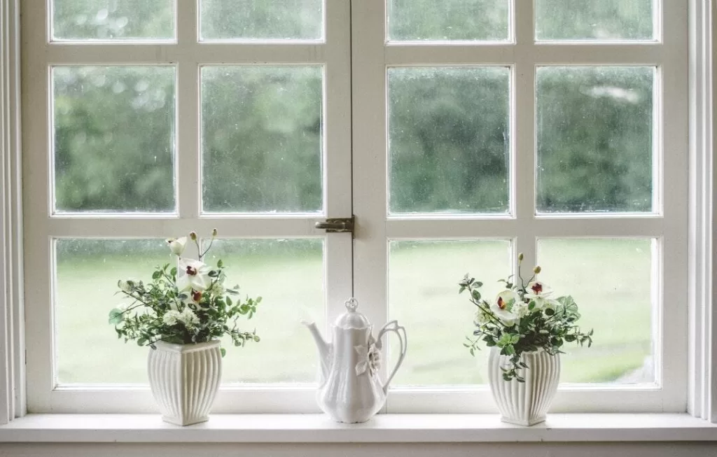 A cozy windowsill featuring two vases with fresh white flowers and a classic white teapot, overlooking a lush green garden through rain-speckled windows.