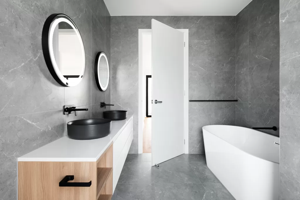Contemporary bathroom featuring grey stone tiles, dual black basin sinks with wall-mounted faucets, round illuminated mirrors, and a freestanding white bathtub.