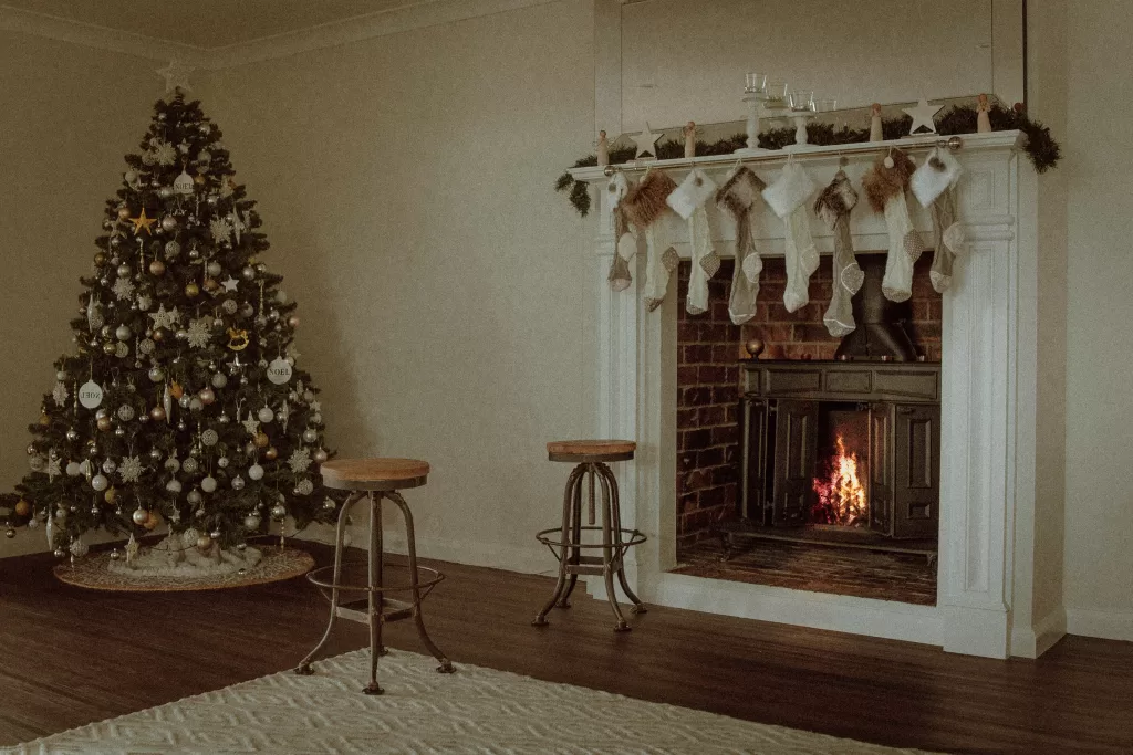 Vintage-styled living room with a traditional Christmas tree adorned with white and golden ornaments, stockings hanging on a classic white mantelpiece above a brick fireplace with a roaring fire, creating a nostalgic holiday atmosphere