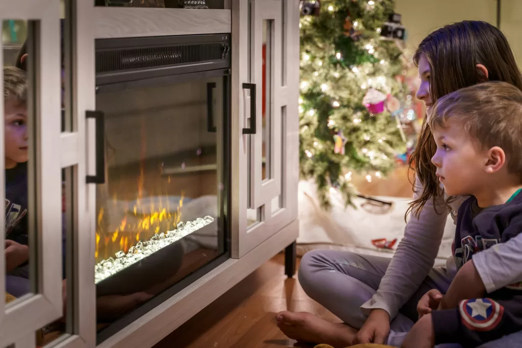 Two young children sitting on the floor gazing at an electric fireplace, with reflections visible in the glass, and a brightly lit Christmas tree in the background, indicating a cozy holiday scene at home.