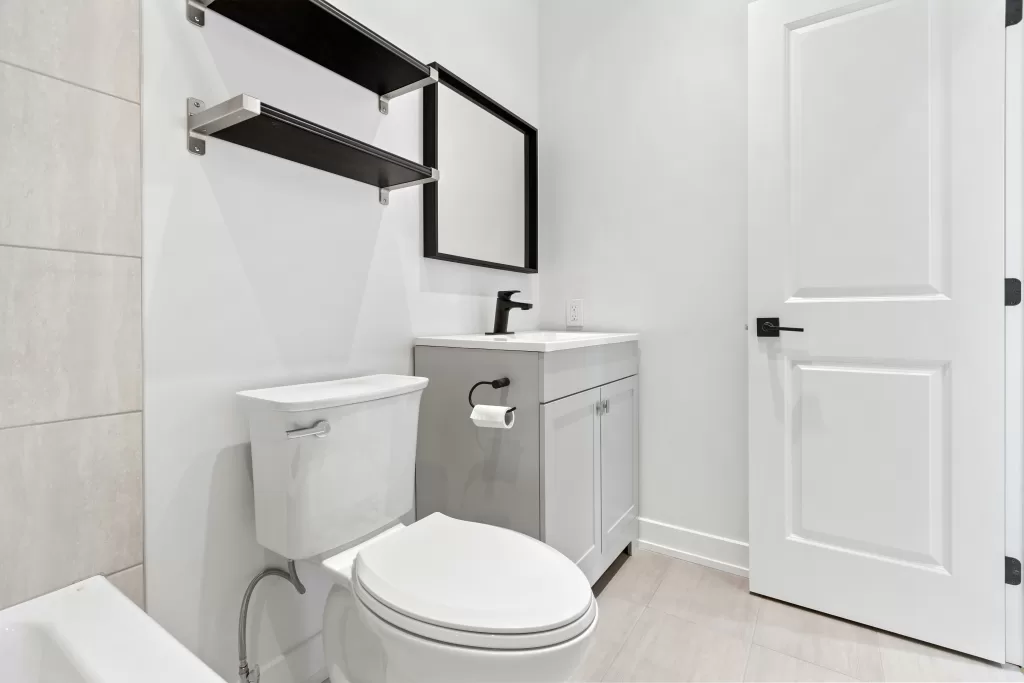 A contemporary bathroom setup with an upflush toilet, white vanity cabinet, and a mounted shelf and mirror above, showcasing a clean and simple design.